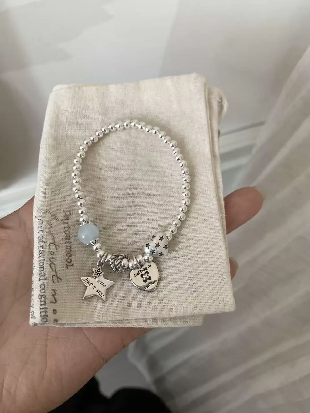 Original seller photo before AI processing — silver charm bracelet held in hand over a cloth jewelry pouch in window light