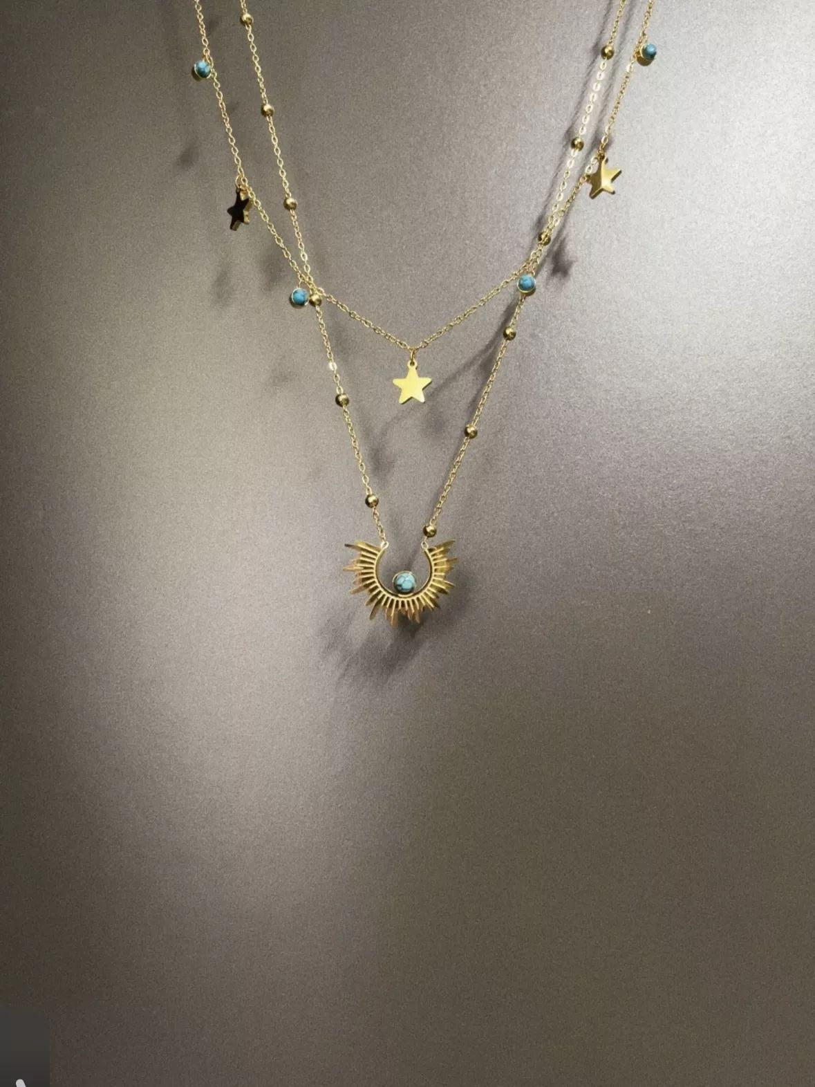 Source supplier photo of a layered gold necklace with turquoise beads and star charms, shot against a gray wall with a hard shadow and the top of the chains cropped out of frame