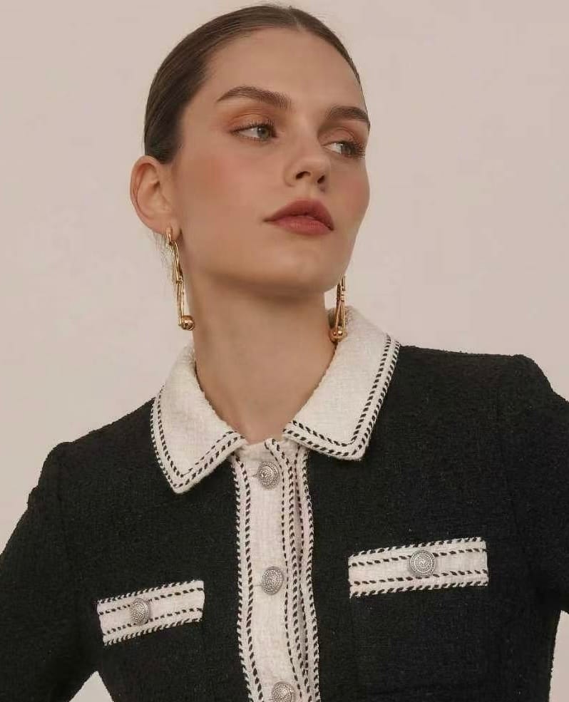 Source product photo: a model in a black tweed jacket with cream contrast collar, braid trim and silver buttons, shot three-quarter against a neutral backdrop with the face turned away from camera.