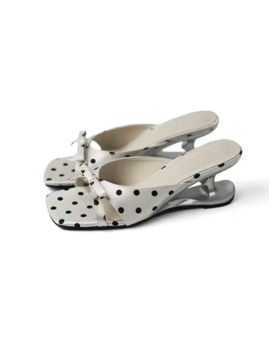 A pair of white polka-dot mules with a bow and a silver sculpted wedge heel, photographed on white