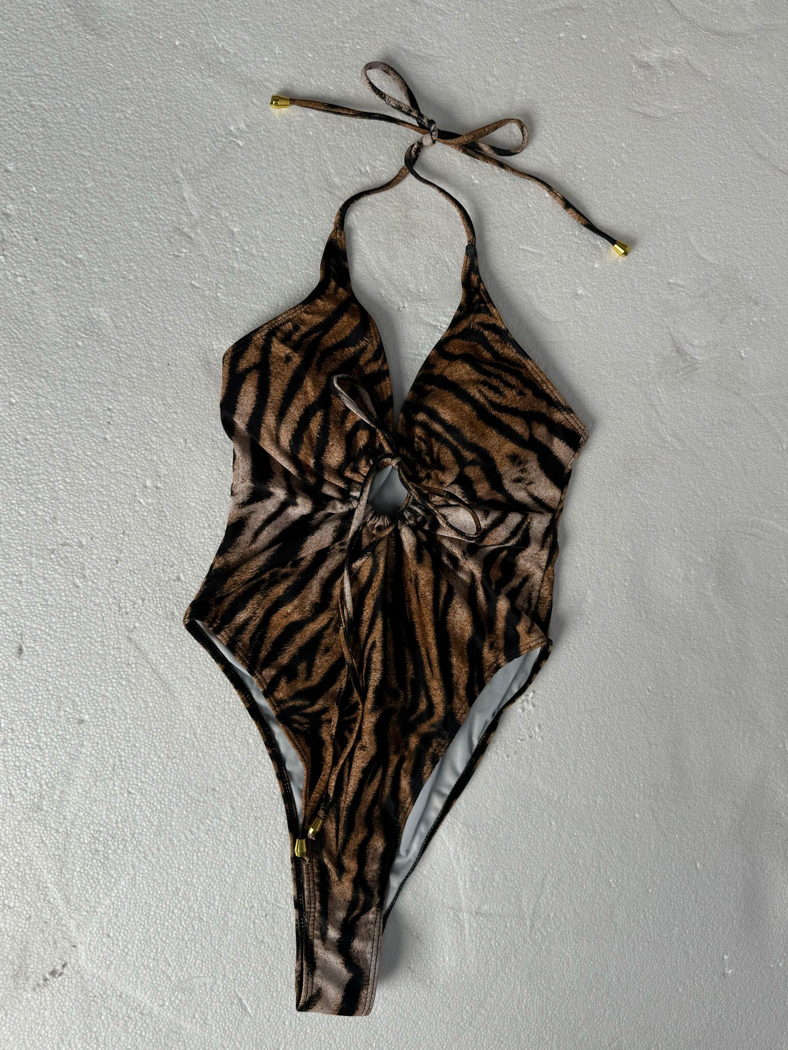 Tiger-print halter one-piece swimsuit raw flat-lay on grey concrete with wrinkled straps and unstructured cup shape — typical supplier file before AI ghost mannequin