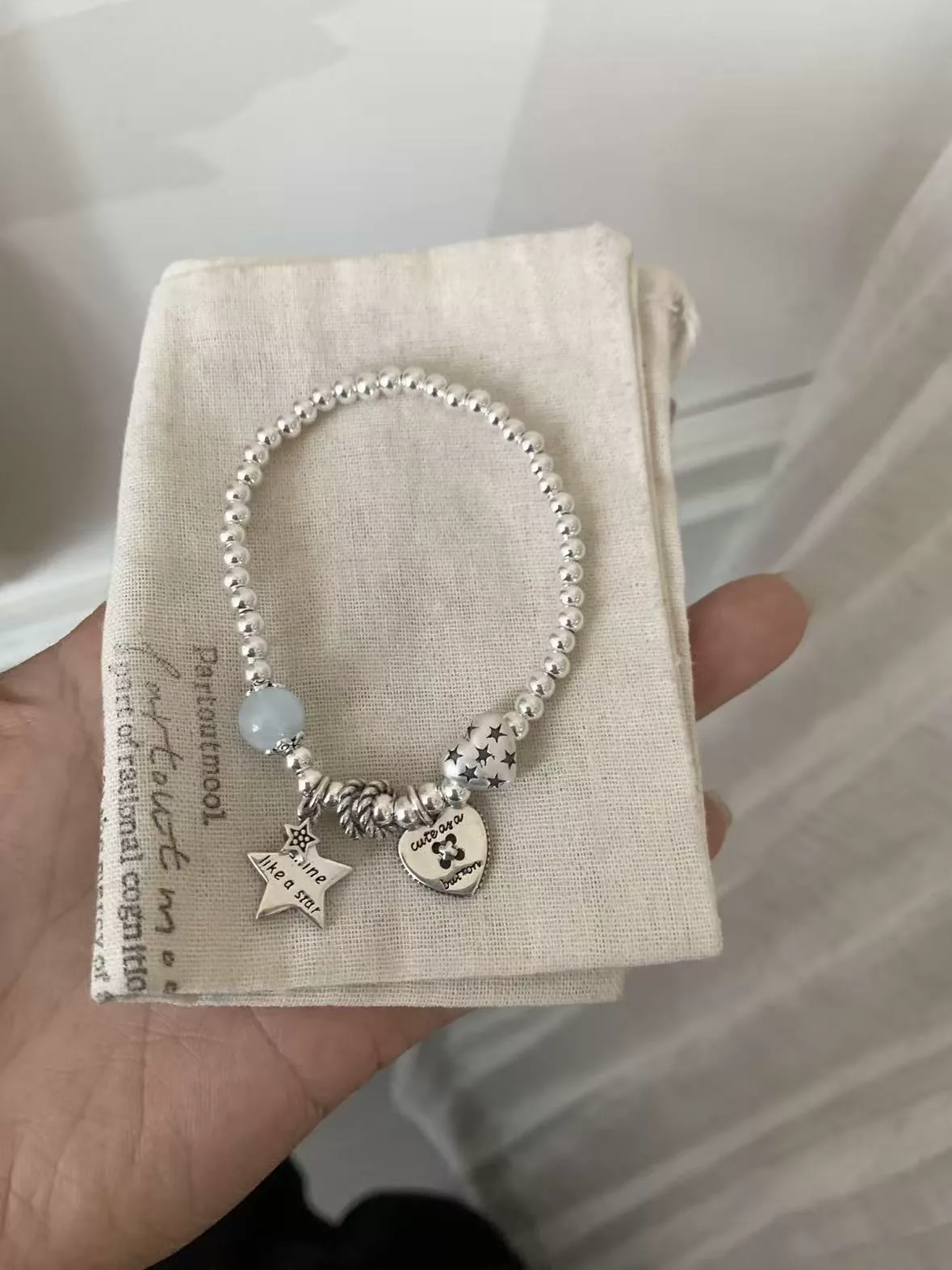 Original seller photo before AI processing — silver charm bracelet held in hand over a cloth jewelry pouch in window light