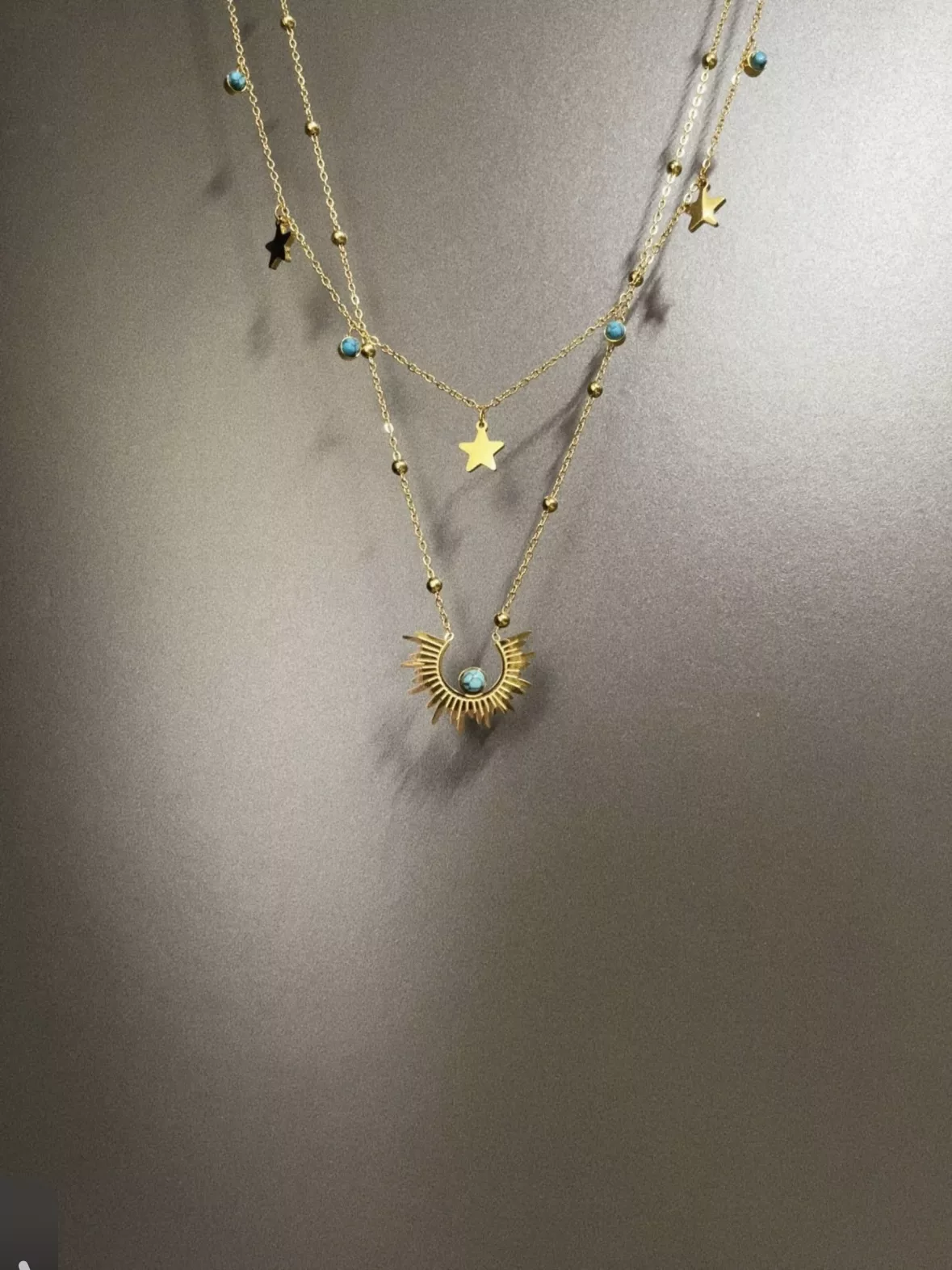 Source supplier photo of a layered gold necklace with turquoise beads and star charms, shot against a gray wall with a hard shadow and the top of the chains cropped out of frame