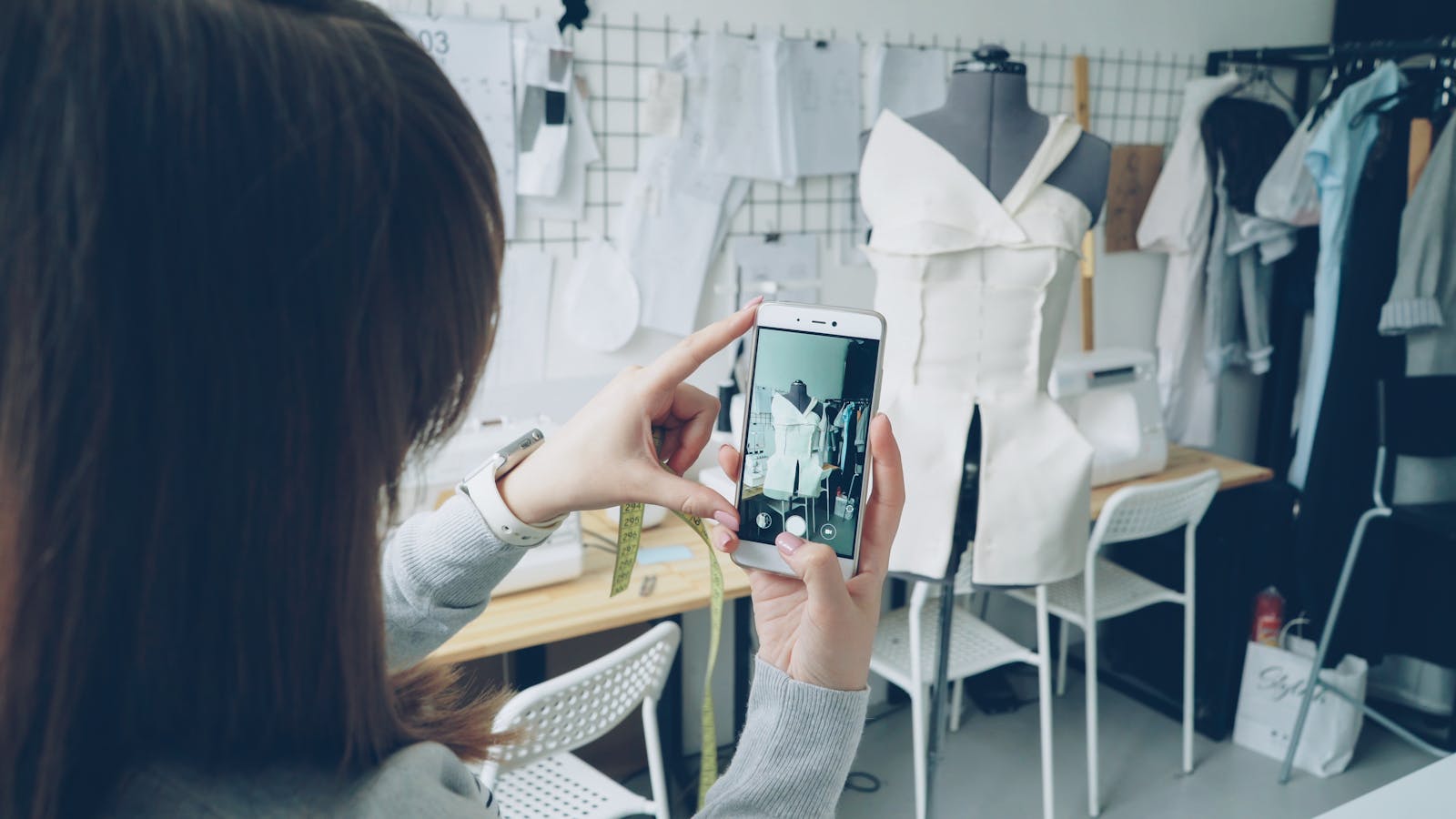 A person photographing a garment on a dress-form mannequin in a studio, framing the shot on a phone — shooting clothing on a mannequin for product photos